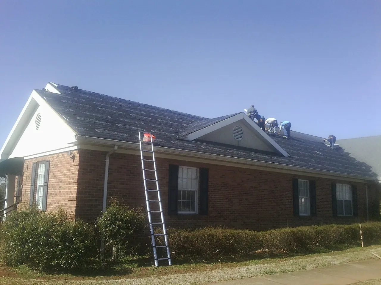 Crew tearing shingles off a brick ranch roof with a ladder set — Metal Roof Repair work by our crew in March Air Reserve Base