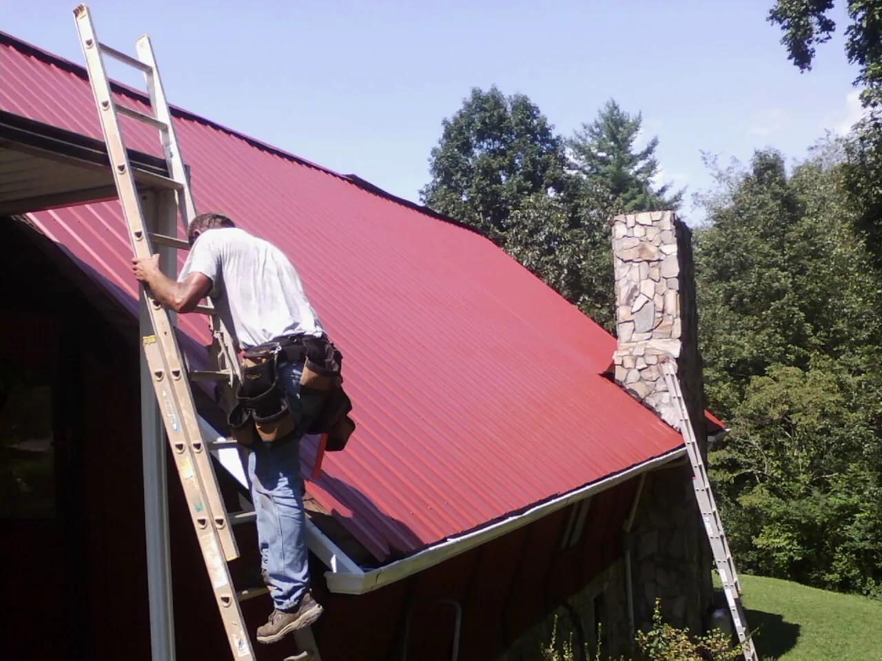 Roofer on a ladder beside a red metal roof and stone chimney — Soffit & Fascia Repair by Roofing Brothers in March Air Reserve Base